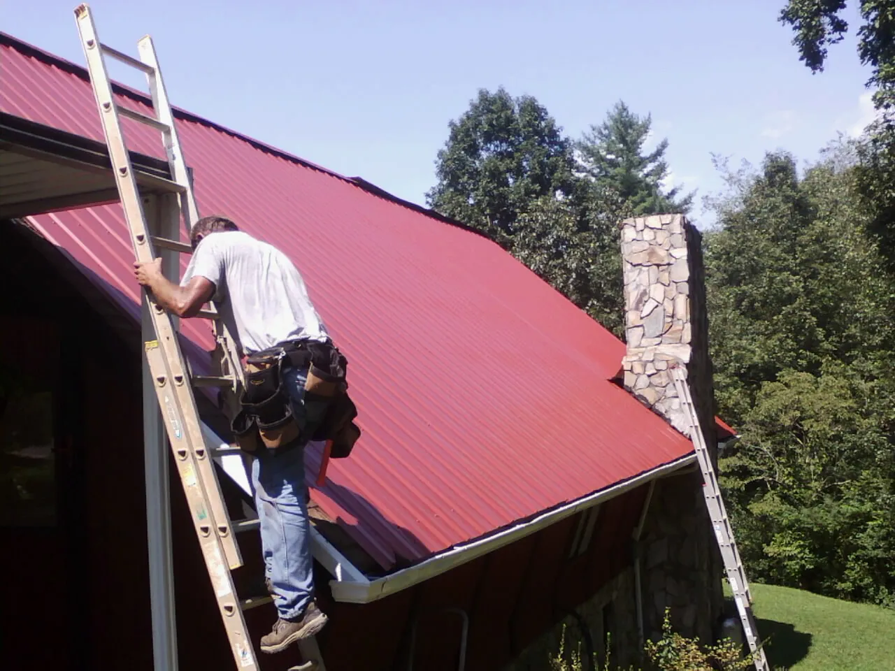 Roofer on a ladder beside a red metal roof and stone chimney — Soffit & Fascia Repair by Roofing Brothers in Lincolnville